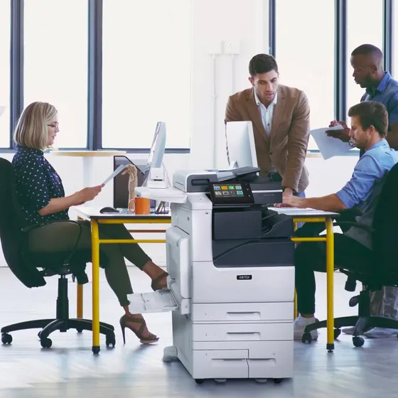 A Xerox C7100 series multifunction printer in a working office, with staff collecting and reviewing documents around it
